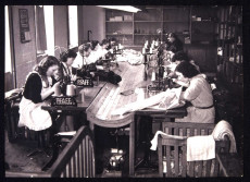 12 couturières sont au travail de part et d'autre d'une grande table dont le milieu est incurvé. Des bobines de fil sont à portée de leurs mains.