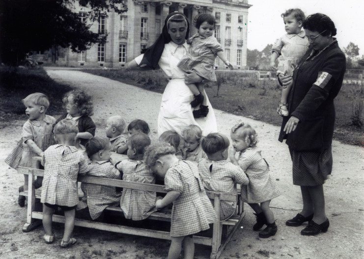 Léa Vion casquée porte un enfant dans ses bras. Une autre femme est en habit de nourrice et porte un voile de nonne. Elle a également un enfant dans les bras. Au sol, dans la cour du château de Bénouville, 12 enfants sont attablés sur des bancs très serrés. Les enfants portent une tenue uniforme.