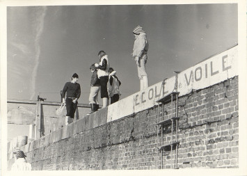 Des jeunes marchant sur la jetée du port, une banderole "école de voile" est accrochée sur le mur de la jetée.