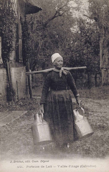 Une femme en tenue traditionnelle porte deux bidons de lait à l'aide d'un portant reposant sur ses épaules.