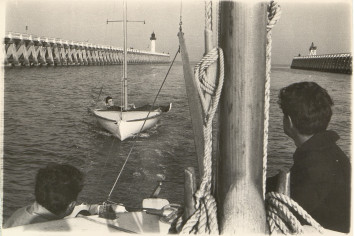 Au premier plan et sur un premier bateau : deux jeunes hommes de dos. Au deuxième plan : un deuxième bateau se fait trainer par le premier, avec un jeune homme à son bord.