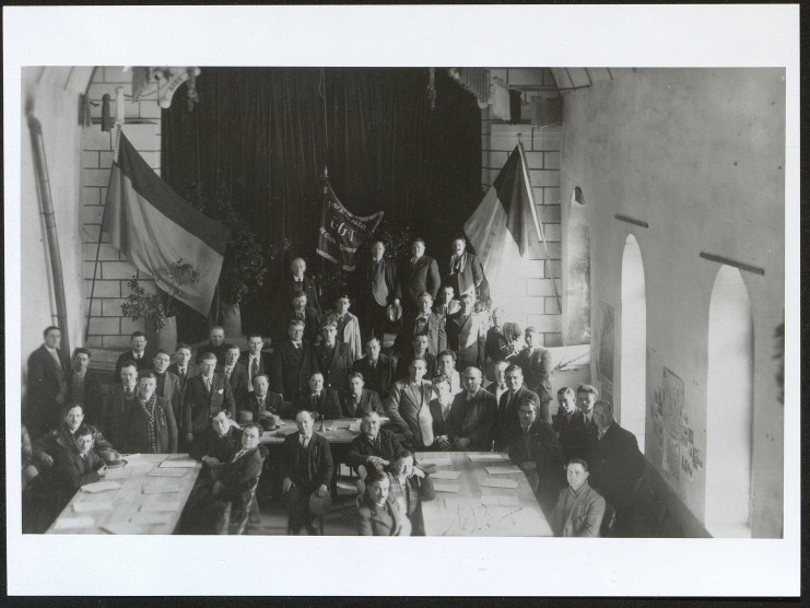 Photographie d'un groupe de membres du syndicat réunis dans une salle et posant, assis ou debout, devant des drapeaux, notamment le drapeau français et celui du syndicat CGT des métaux de Caen et ses environs