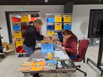 Mise en place de la braderie par deux archivistes. Les livres sont déposés dans des cartons.