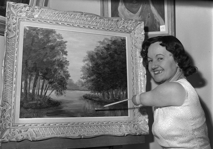 Denise Legrix souriante tient un peint avec son bras gauche qui n'a pas de main. Son pinceau semble être au bout de son bras comme s'il en est le prolongement.