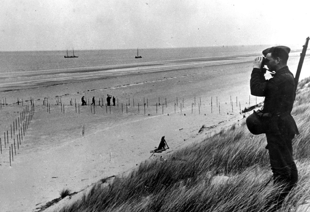 Un soldat allemand scrute avec des jumelles l'horizon sur une  plage calvadosienne comportant des éléments défensifs.