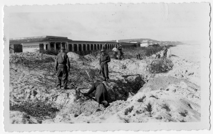 La photographie représente les Planches de Deauville défigurées par les traces des combats ainsi que des barbelés. Des soldats sont présents sur les lieux.