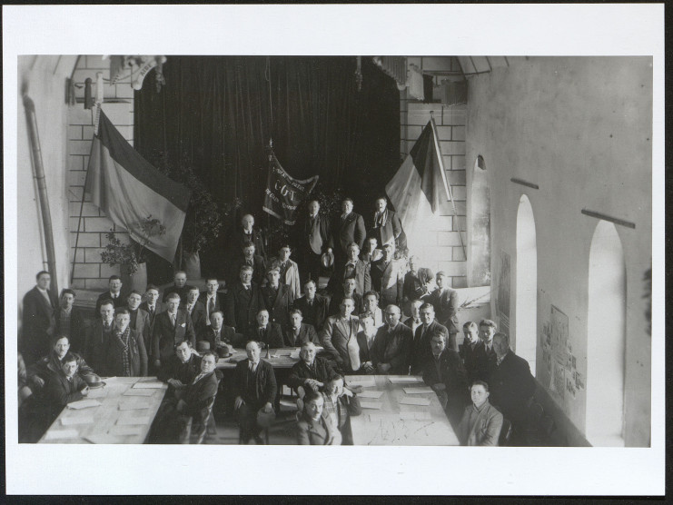 Photographie d'un groupe de membres du syndicat réunis dans une salle et posant, assis ou debout, devant des drapeaux, notamment le drapeau français et celui du syndicat CGT des métaux de Caen et ses environs